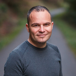 Professional headshot of a male CTO and software architect in smart casual attire, thoughtful expression, modern tech workspace with multiple monitors, natural lighting