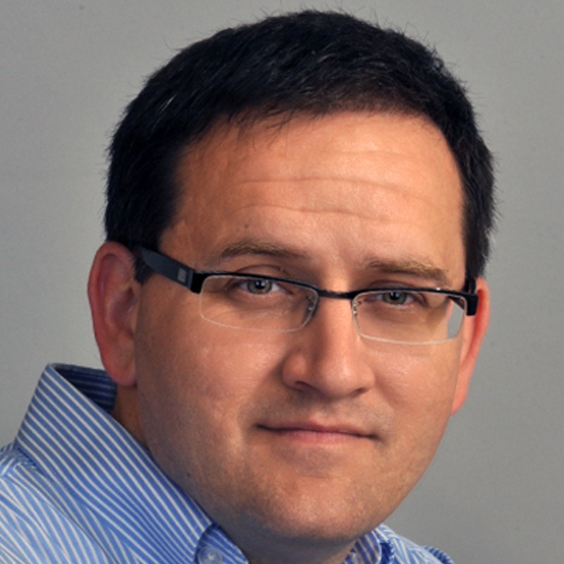 Professional headshot of a male ESG expert in his 40s wearing a dark business suit, confident expression, European office background with EU flags, warm studio lighting